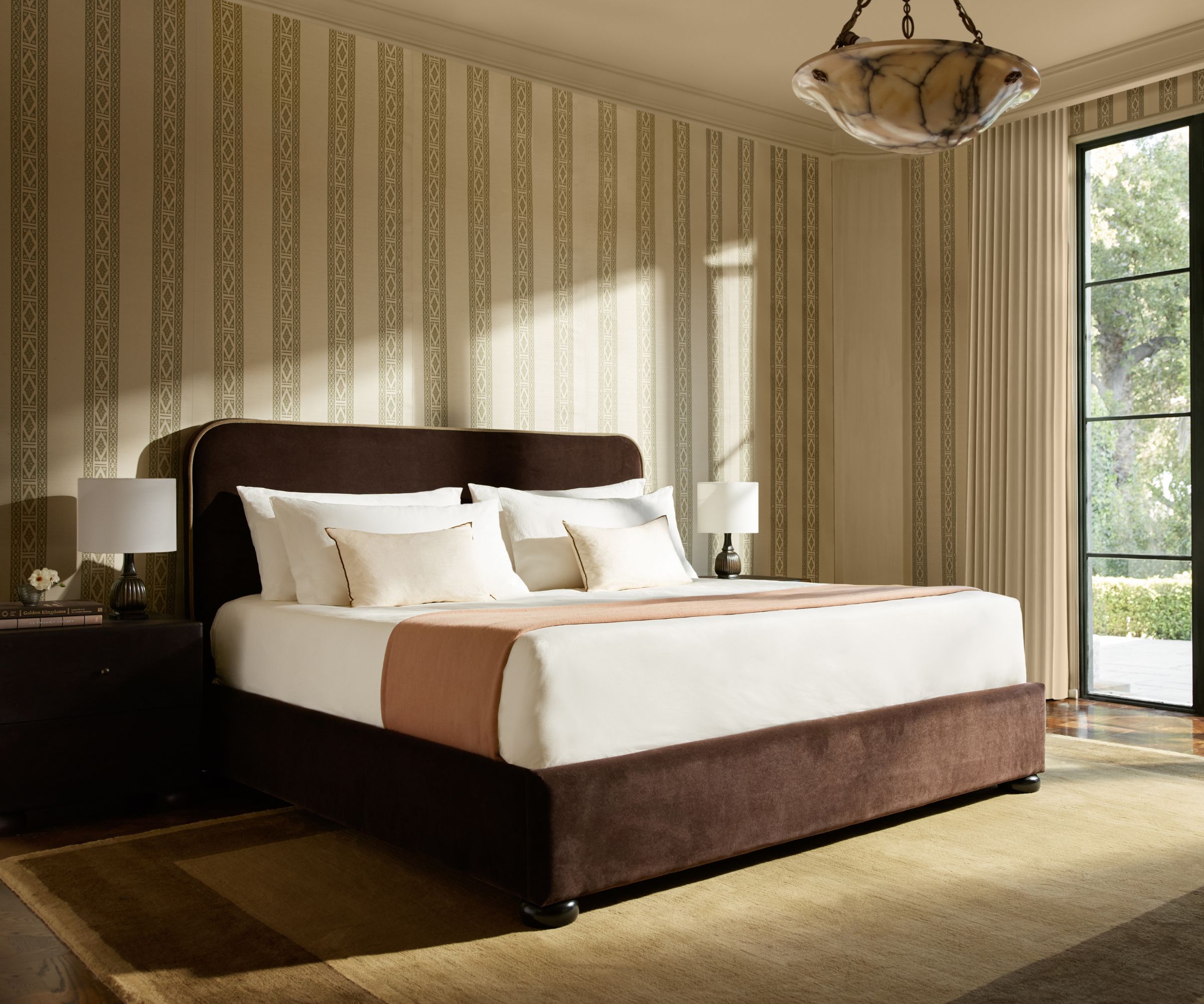 A neutral bedroom with stripe wallpaper, a large earth-toned rug, and a brown velvet bed frame