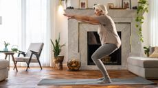 woman sideways on to the camera, performing a squat with arms raised in a living room setting 