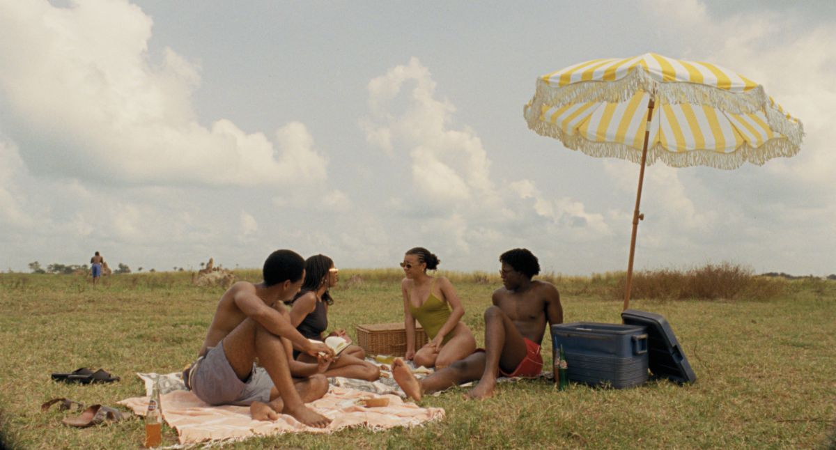 two men and two women sit in bathing suits under an umbrella on picnic blankets in a still from clarissa