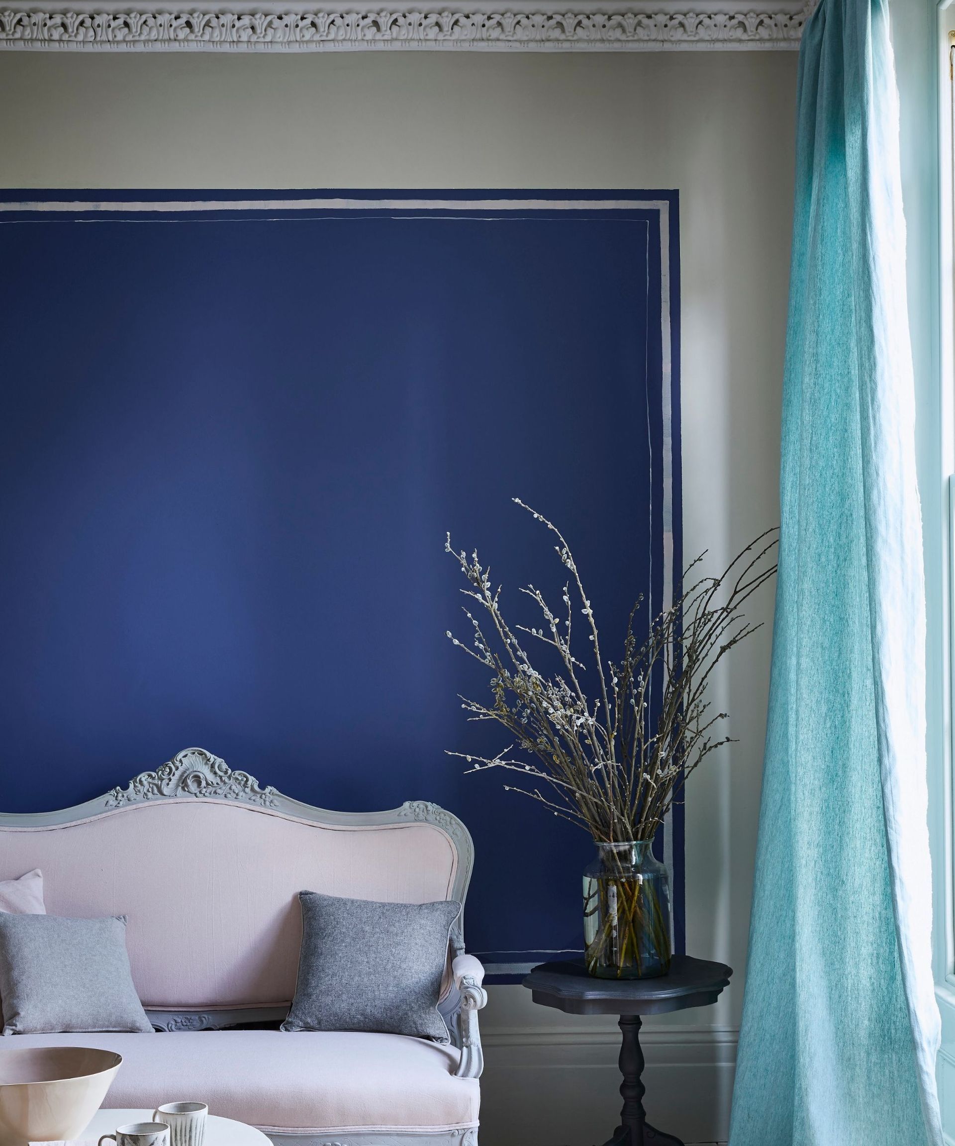 A wall painted in navy blue, there is a pale pink sofa with grey scatter cushions, next to it is a small black side table with flowers in it.