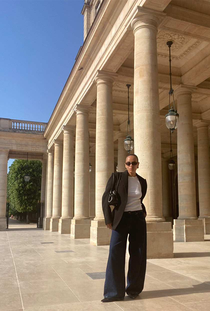 An editor standing at Palais-Royal wearing a black blazer, white tank top, barrel-leg jeans, black pointed flats, a black shoulder bag, and sunglasses.