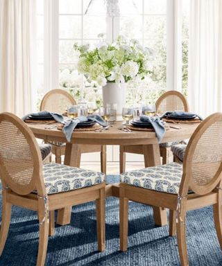 Four rattan chairs around a circular dining table; blue napkins on each place setting; in the middle, a vase filled with white flowers against windows with a garden view.