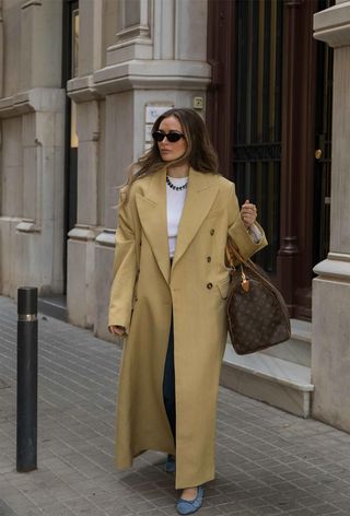 A winter travel outfit is shown in a photo of a woman standing on a sidewalk wearing a long tan coat over a white shirt and straight-leg jeans styled with a silver necklace, denim ballet flats, black sunglasses, and a Louis Vuitton duffle bag