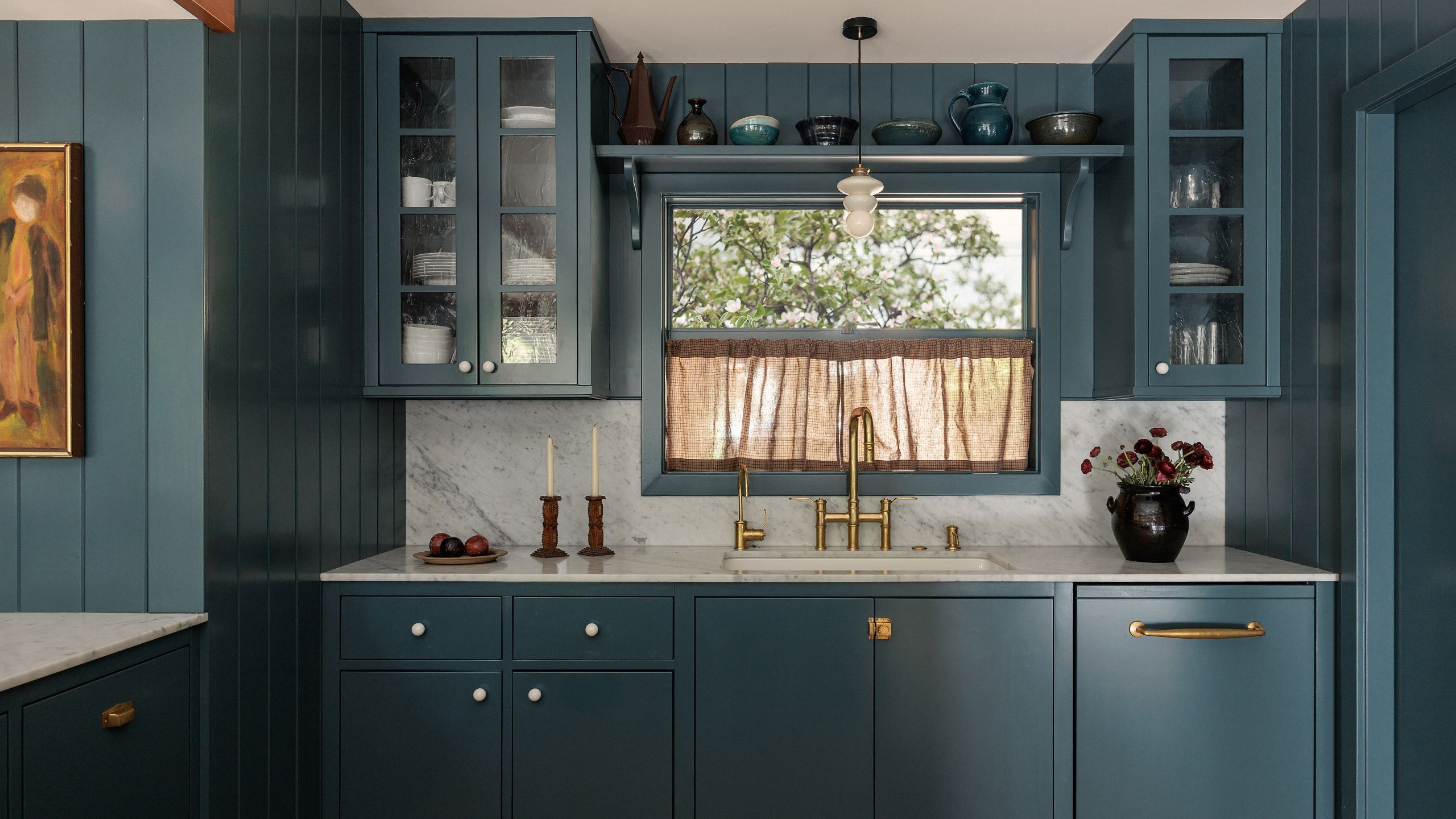 a blue green warm kitchen with wooden beam detail, small window with cafe curtain, glass upper cabinets with an open shelf and paneling