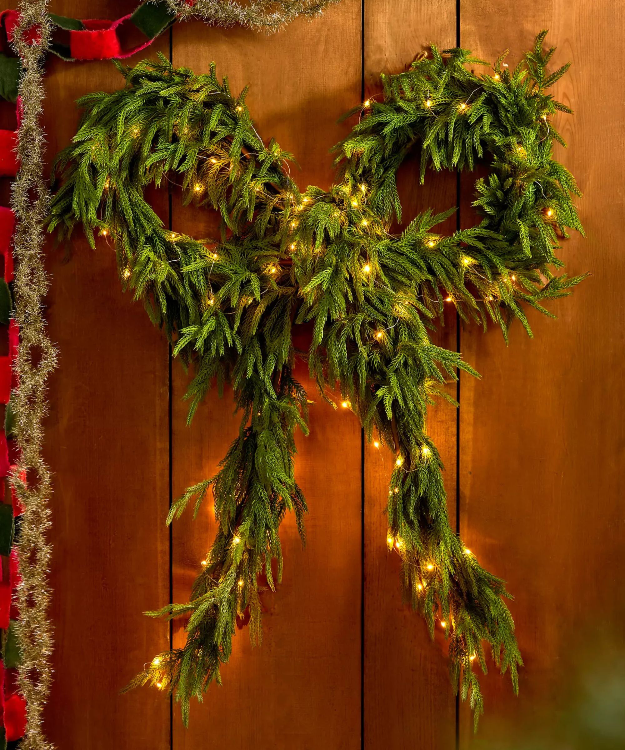 A bow-shaped wreath woven with fairy lights that sits against a wooden wall. Colourful paper chains and silver tinsel are beside it.