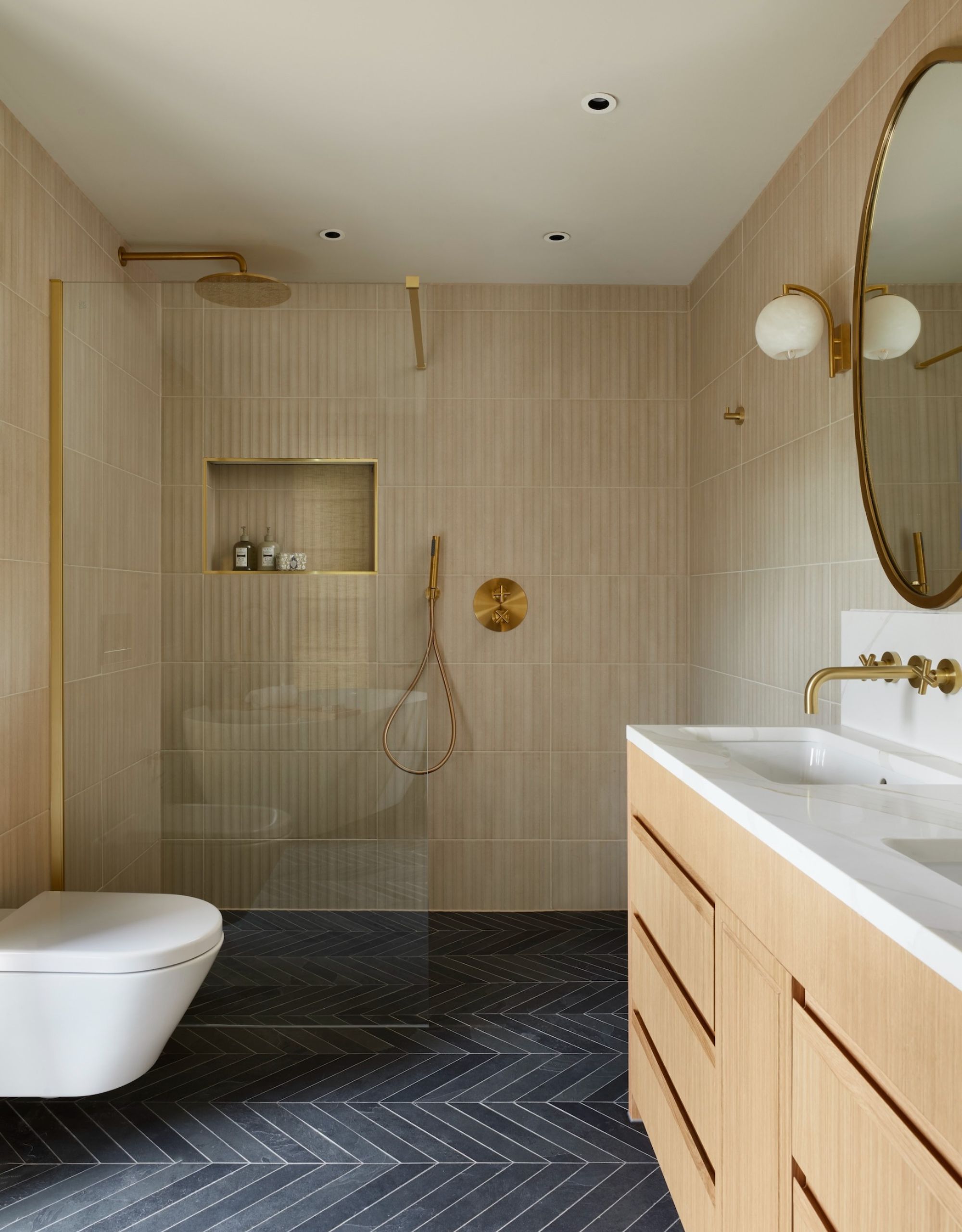 A wide walk in shower shown with a wall-hung wc and double vanity basin. Neutral wall tiles and slate grey flooring in a herringbone layout