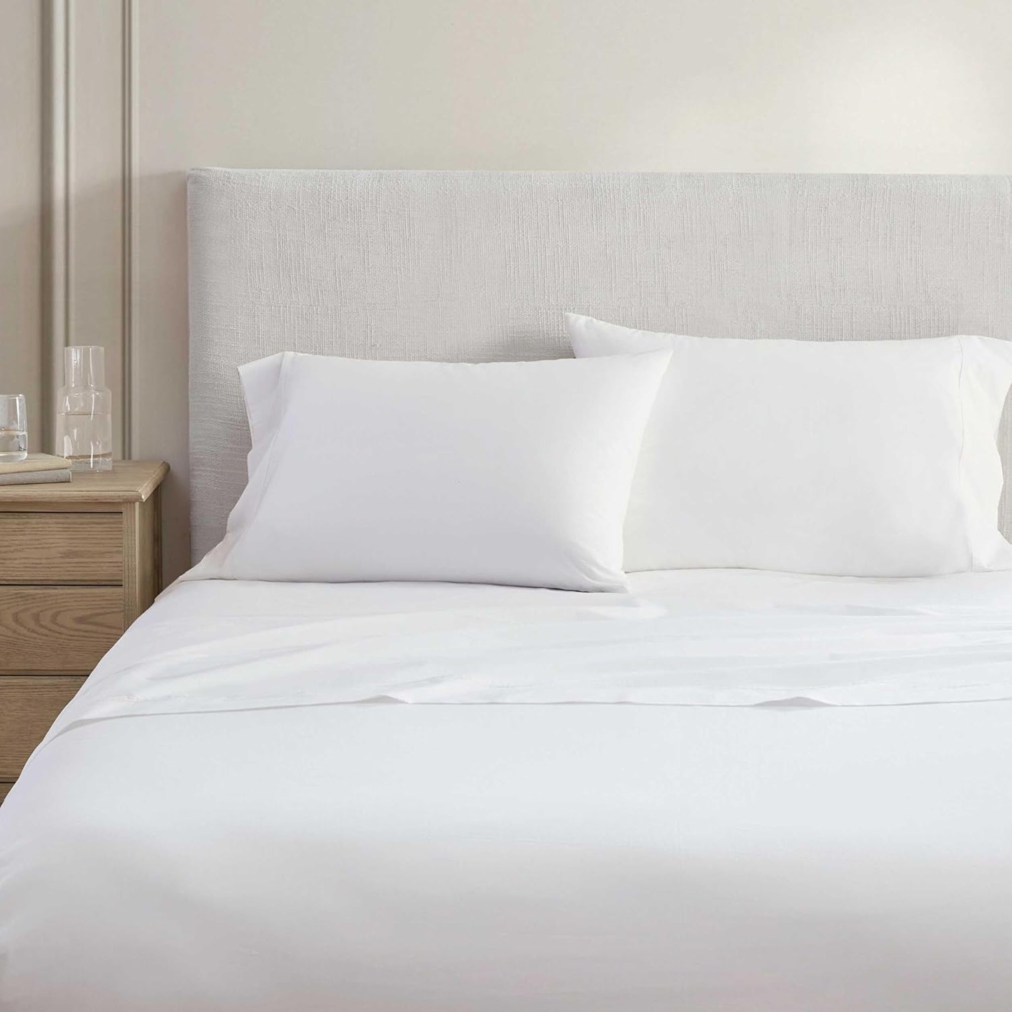 Martha Stewart white Sheet set on a white bed with cream panelled wall behind and light wood nightstand with glassware to the left.