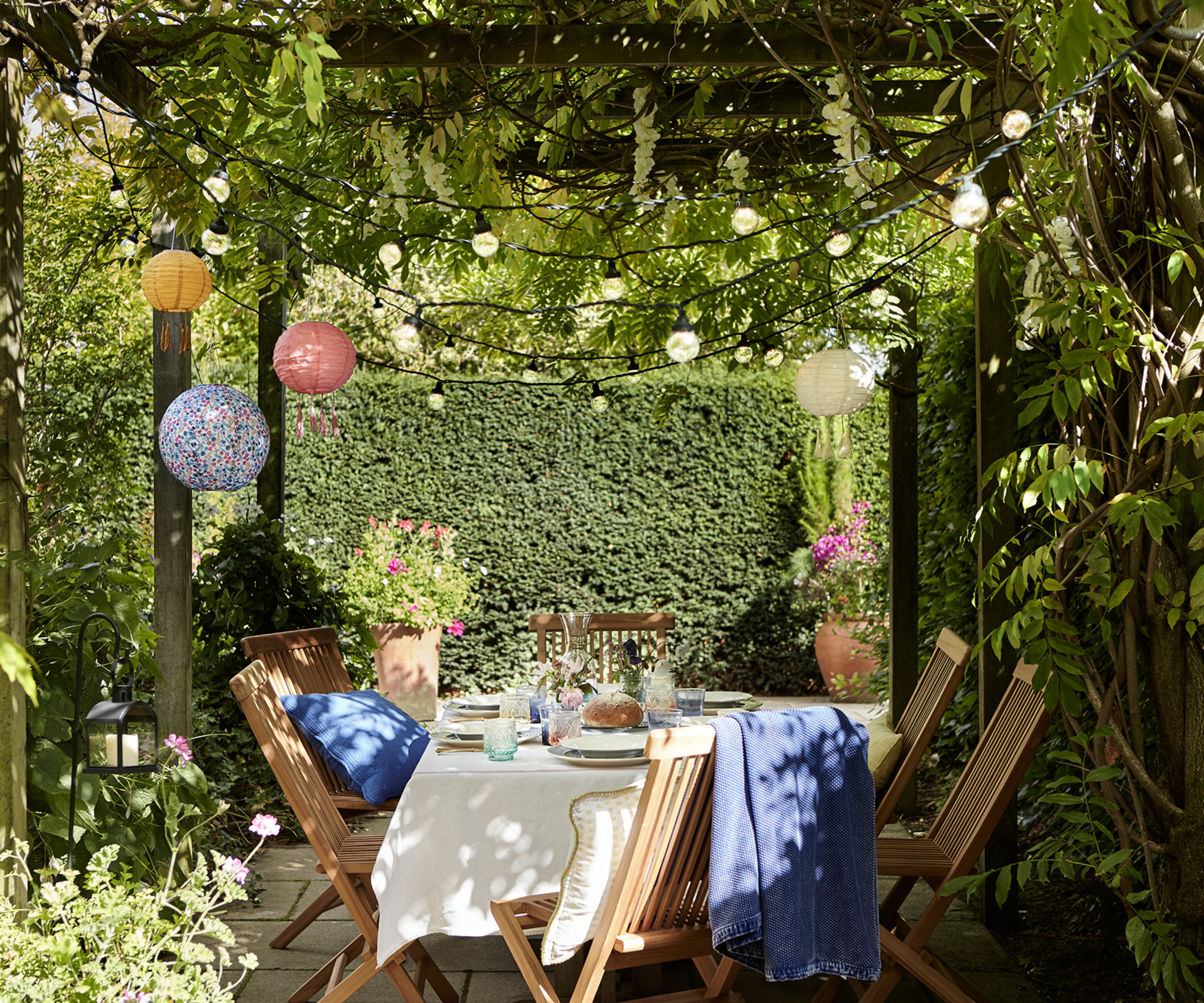 wooden pergola with planting grown over roof and decorative lights hanging over table and chairs