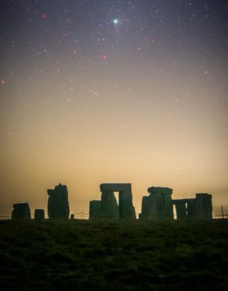 stonehenge pillars are in shadow in the foreground. behind is an orange sky. Far above is a green comet shining in the stars