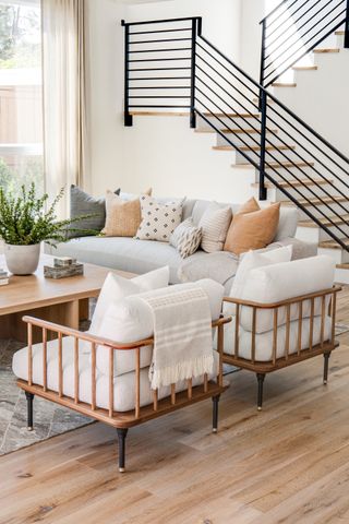 living room with natural color scheme, gray couch, off white armchairs with wood frame, wooden coffee table and floor, black staircase