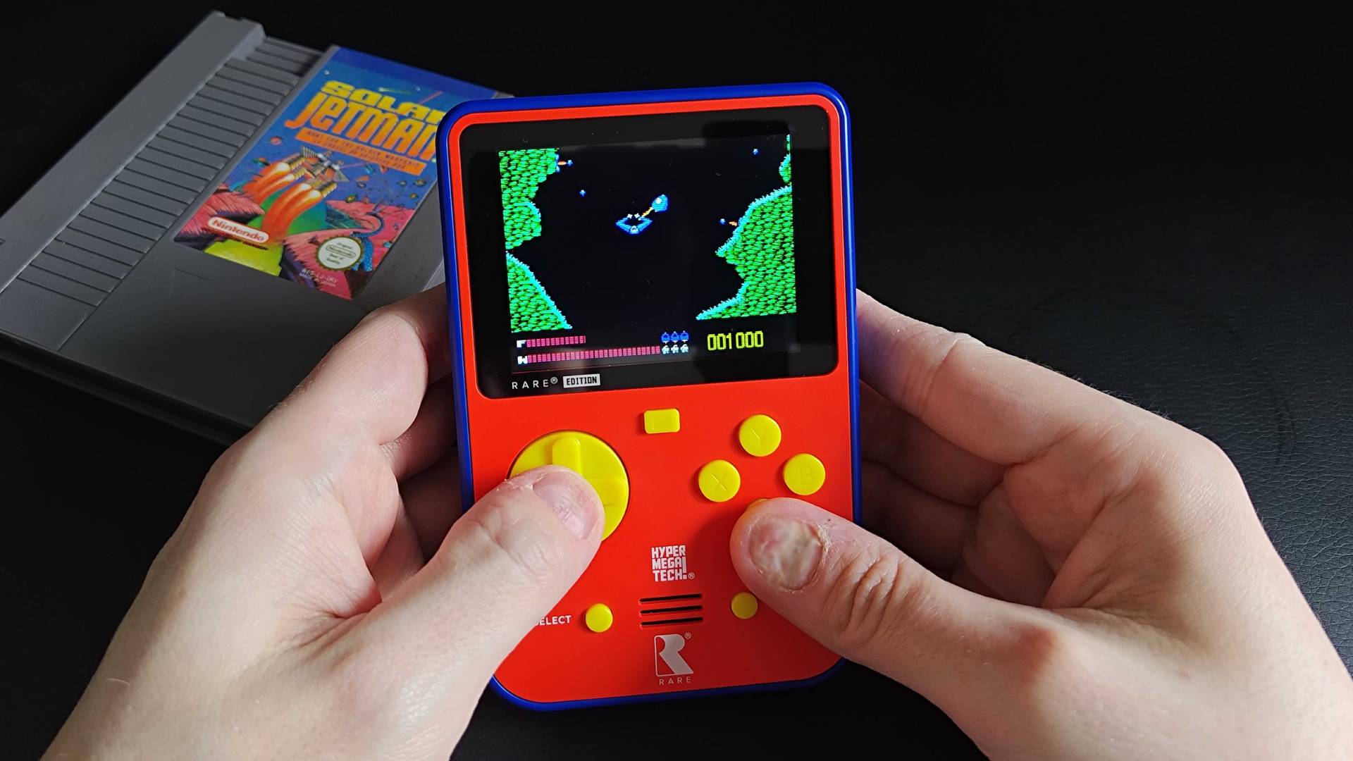 Hands using Super Pocket Rare Edition to play Solar Jetman with NES cartridge on desk in backdrop.