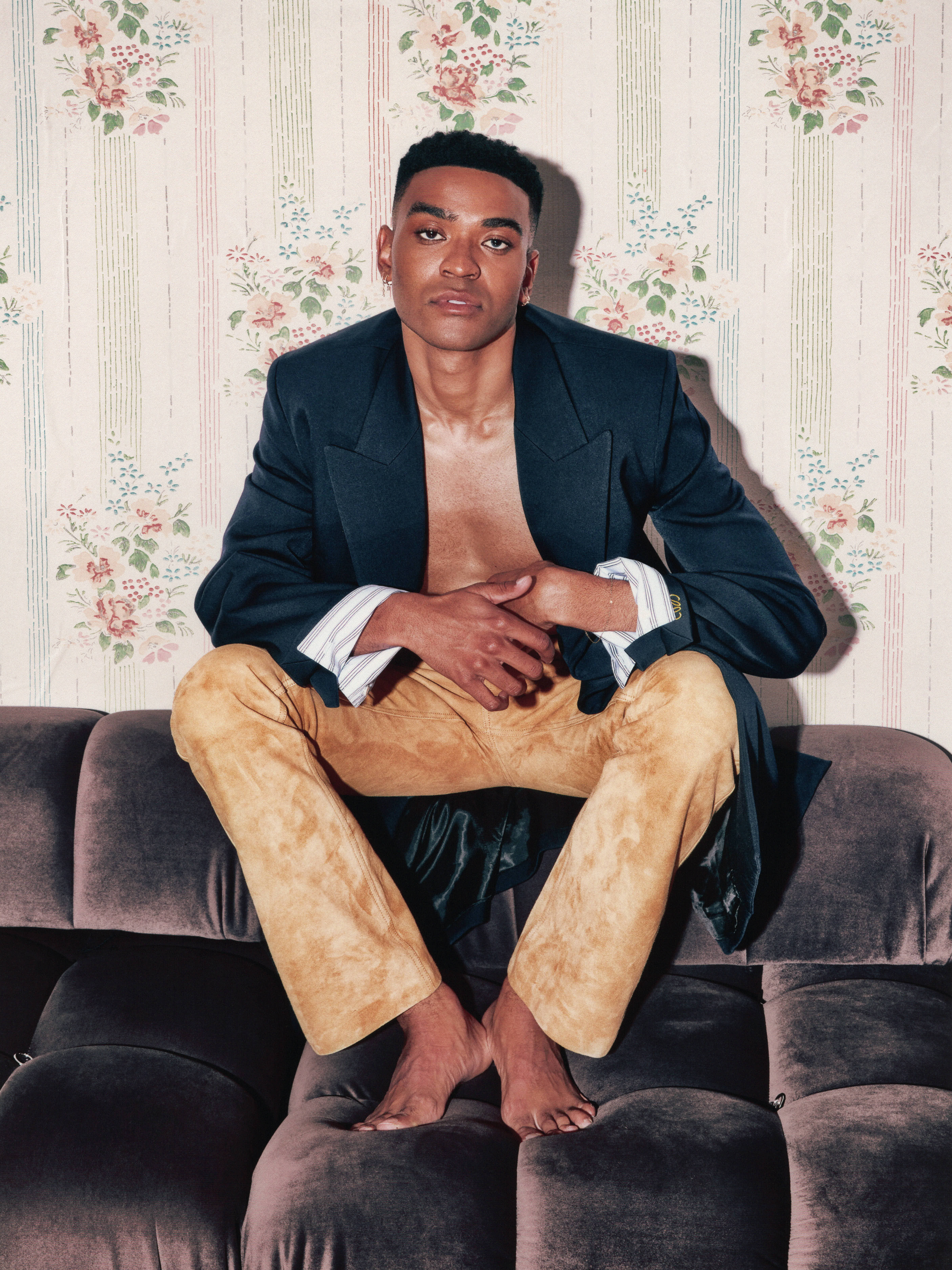 Owen Thiele poses sitting on a grey velvet couch wearing a black jacket with no shirt and tan suede pants