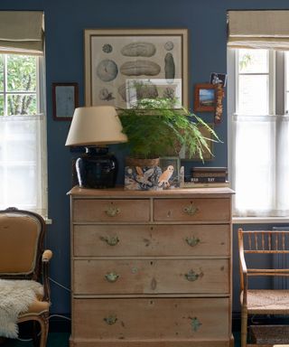 A blue room with an antique dresser, two chairs and artwork