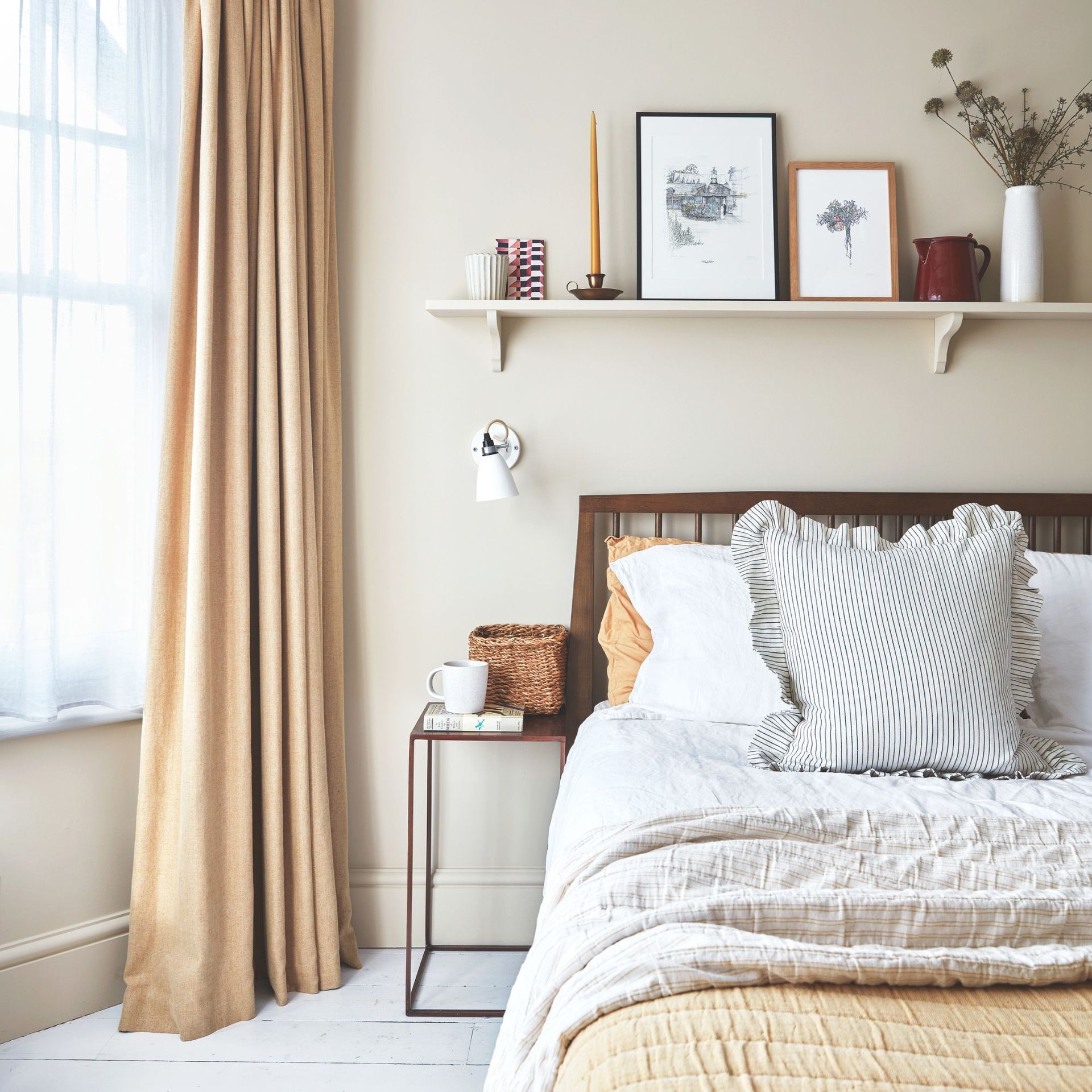 Neutral painted bedroom with cream curtains to the side of a bed with lots of layered bedding on it, and a shelf with trinkets on it above it