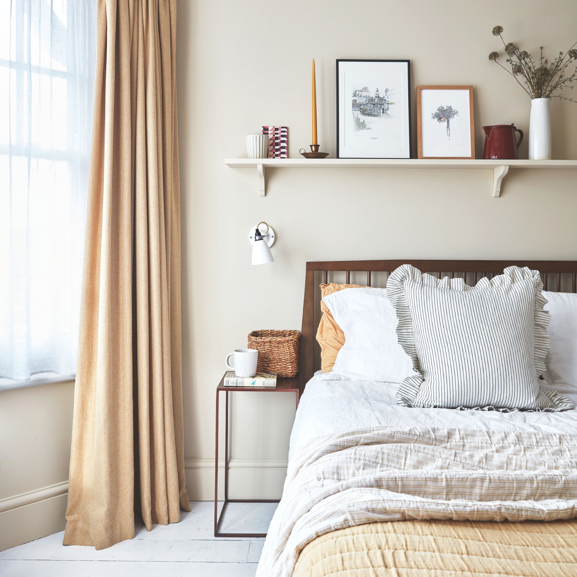 Neutral painted bedroom with cream curtains to the side of a bed with lots of layered bedding on it, and a shelf with trinkets on it above it