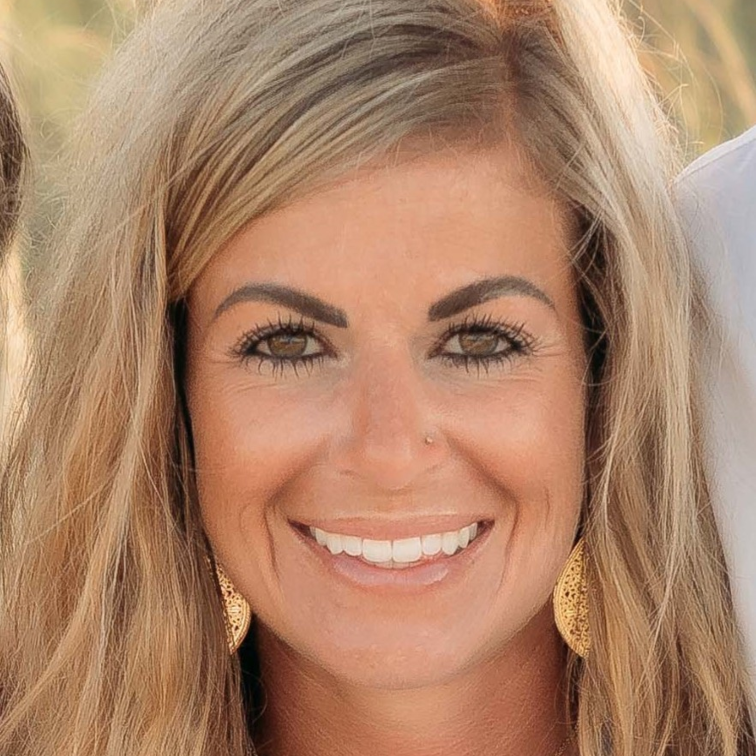 A headshot of a smiling woman with wavy blonde hair, dangling gold earrings and a bright smile