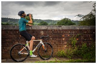 A male cyclist on his bike leans against a wall and drinks from a bottle