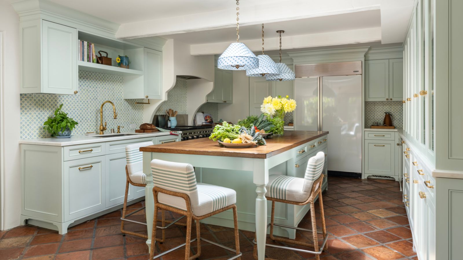 A midimalist kitchen with blue-green cabinets, terracotta floors, and a wooden countertop on the island