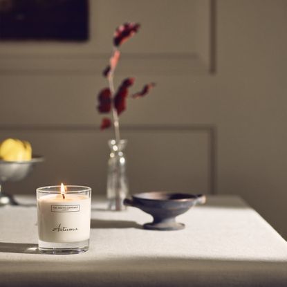 The White Company's lit Autumn signature candle on a dining table with a bowl of fruit and a vase with a single stem in the background