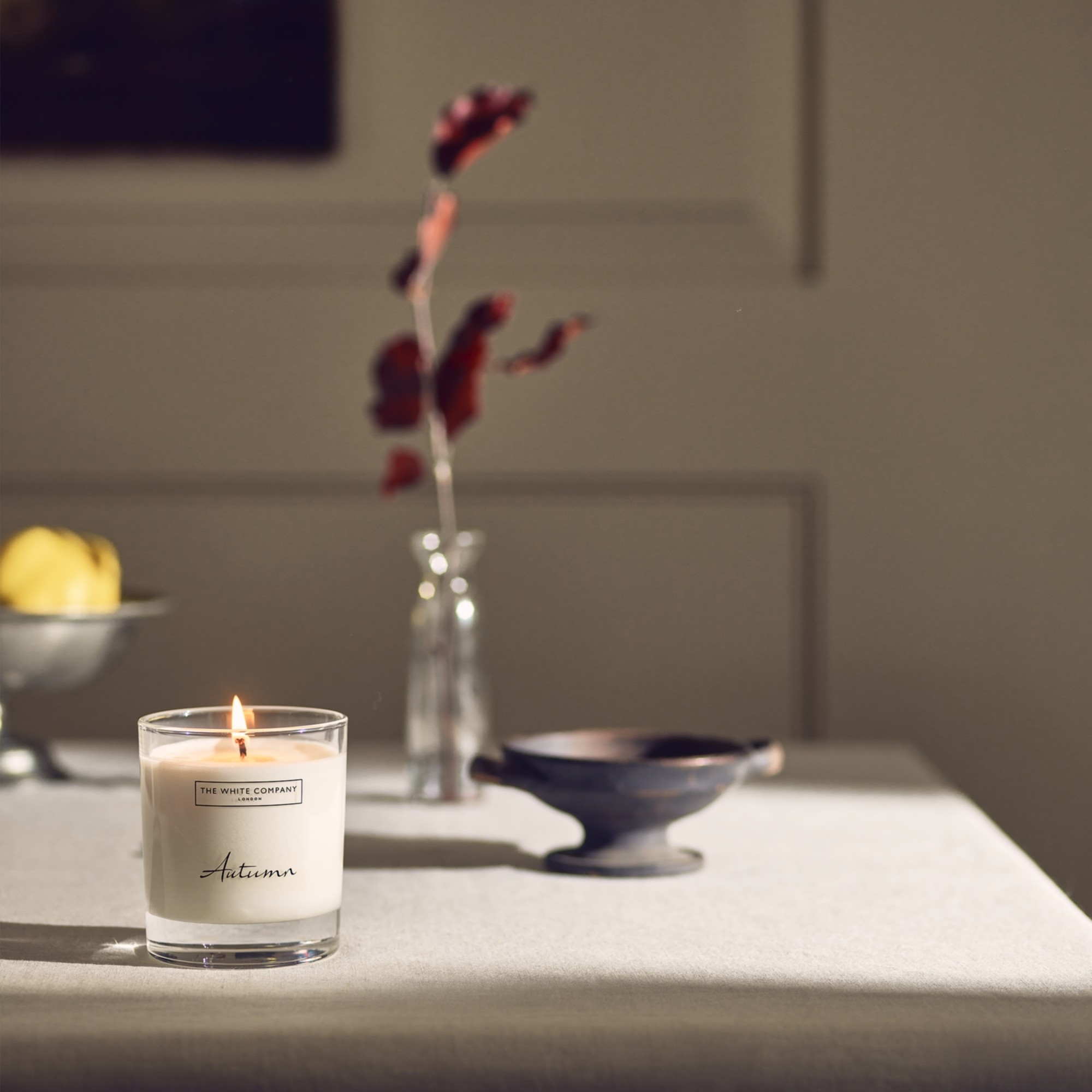 The White Company's lit Autumn signature candle on a dining table with a bowl of fruit and a vase with a single stem in the background
