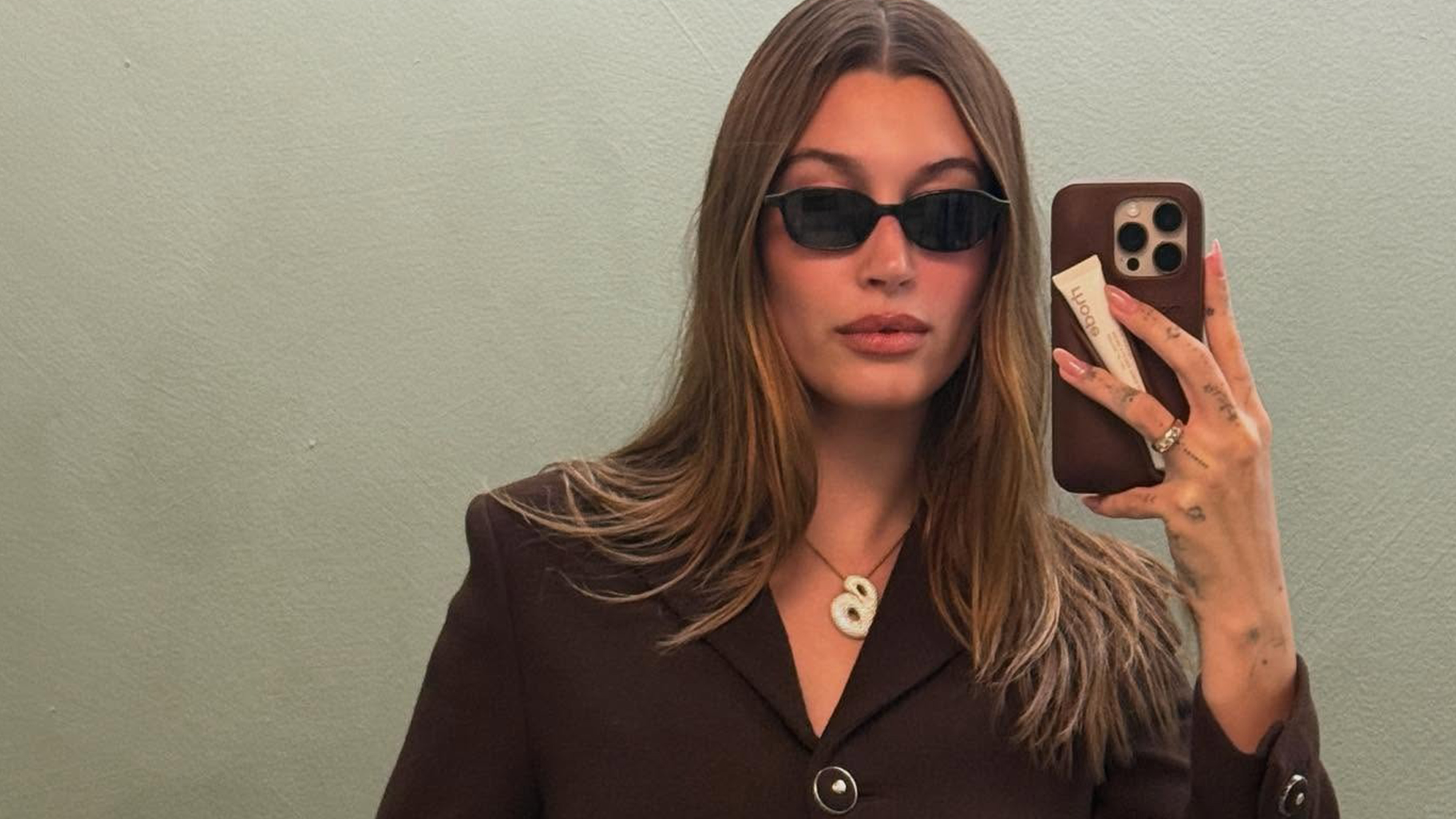 Hailey Bieber wearing sunglasses, a brown blazer, and her B necklace.