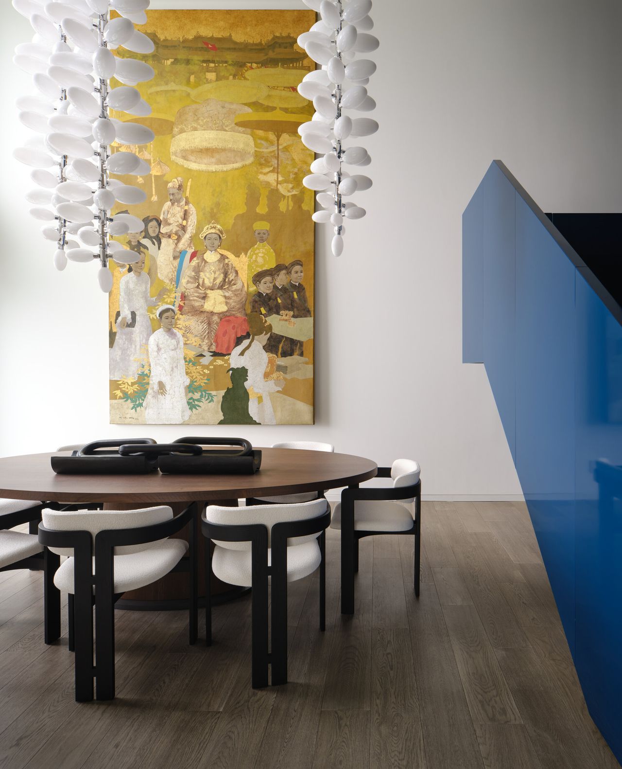 open plan dining room next to blue staircase with round timber dining table and white and timber dining chairs, a large yellow artwork on the wall, and statement pendant lights draping down above the table