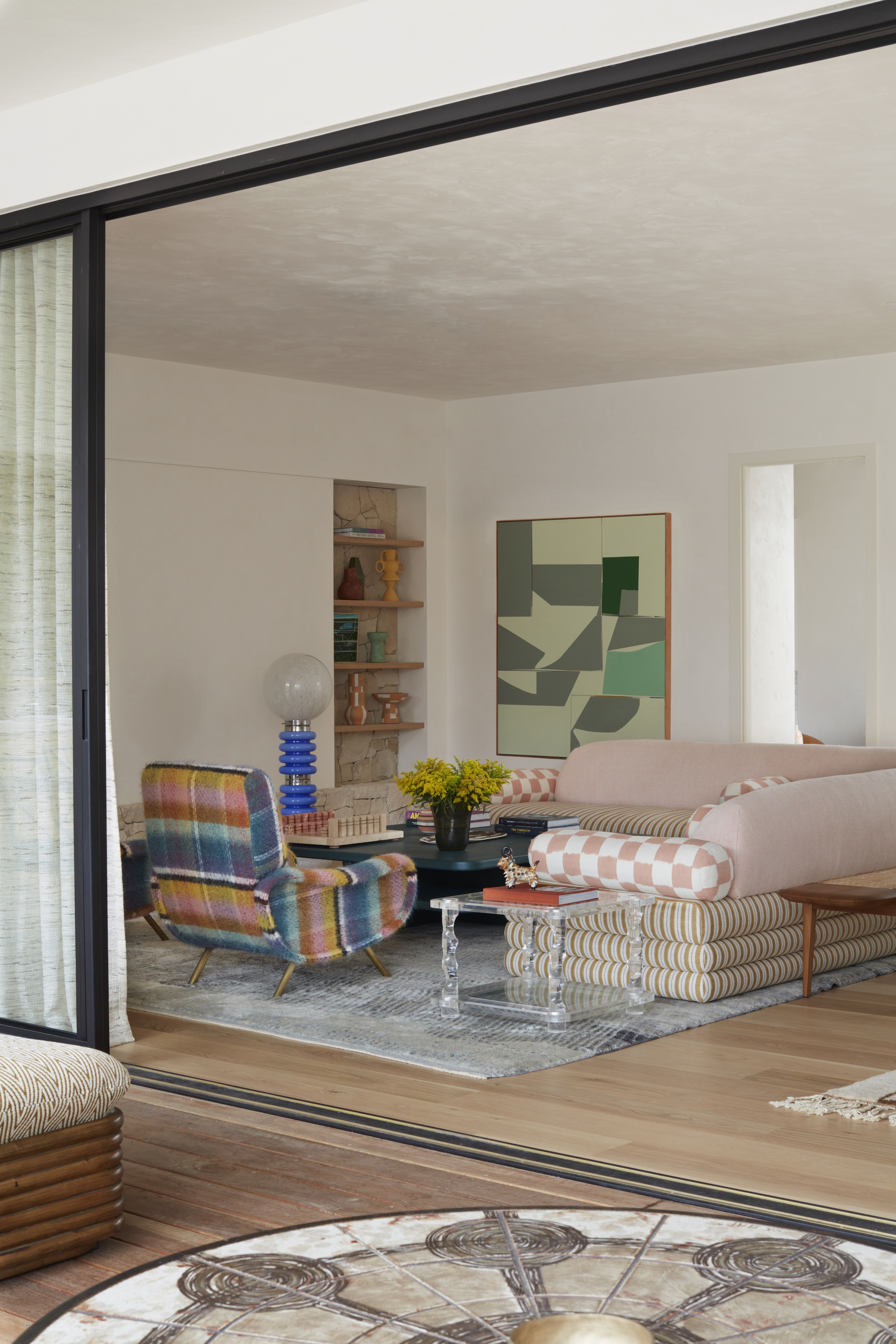 A view into a modern living space from the patio. There is a large corner sofa, to the right and a patterned armchair, both around the coffee table, with an art piece on the far wall.