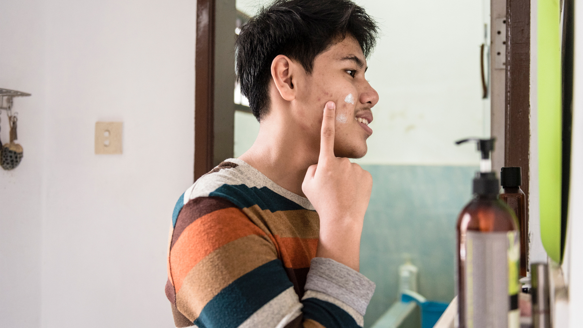 young man applying cream to his facial acne while looking in the mirror