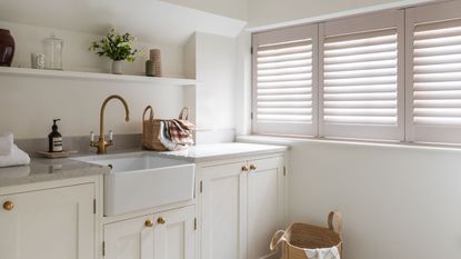 A white laundry room with white cabinets, walls, a sink, and shutters with gold doorknobs and faucets, and marble countertops.