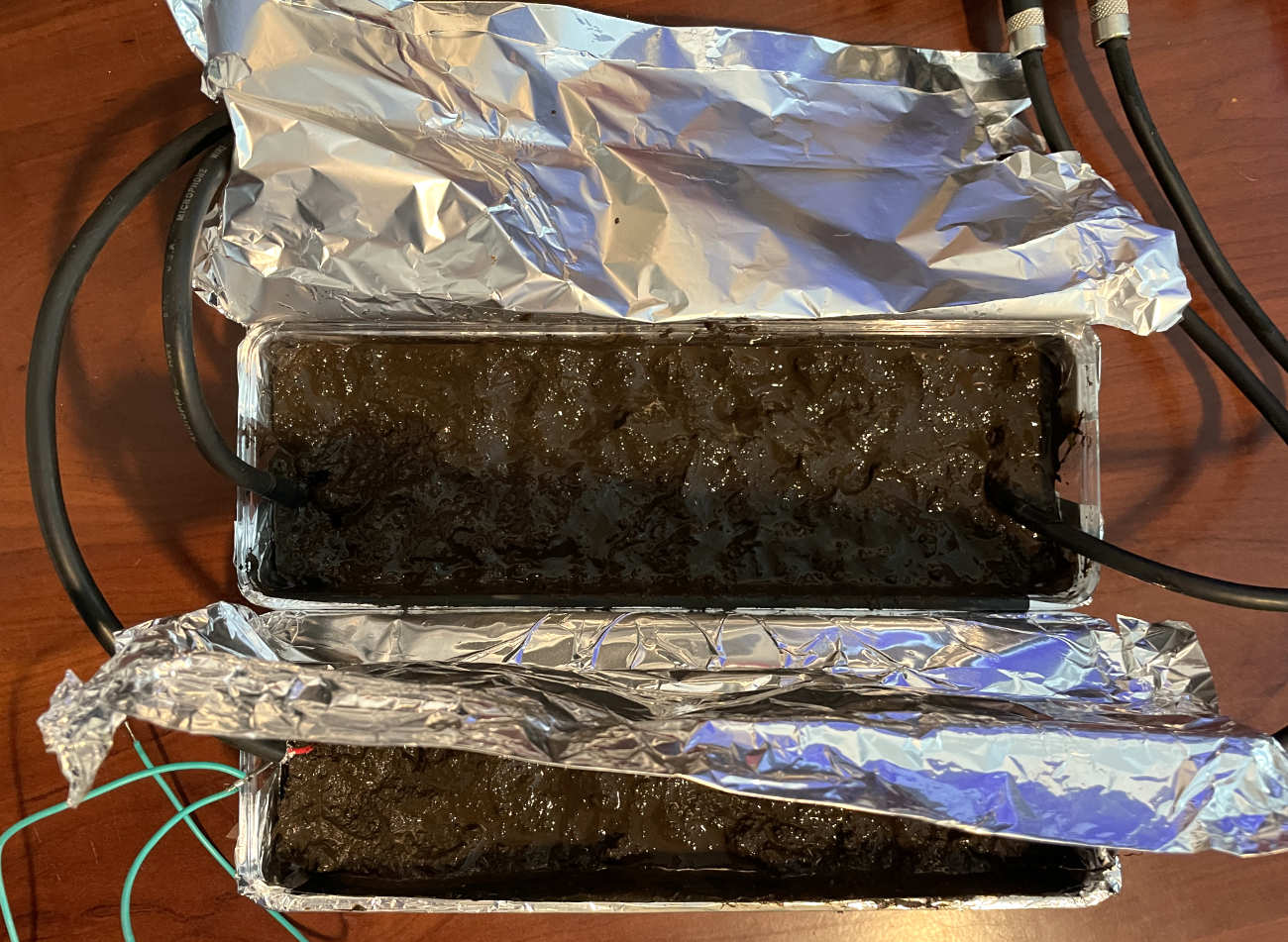 A high-angle shot looking down at two separate aluminium foil containers filled with dark, mud, each acting as a conductor for black audio cables.