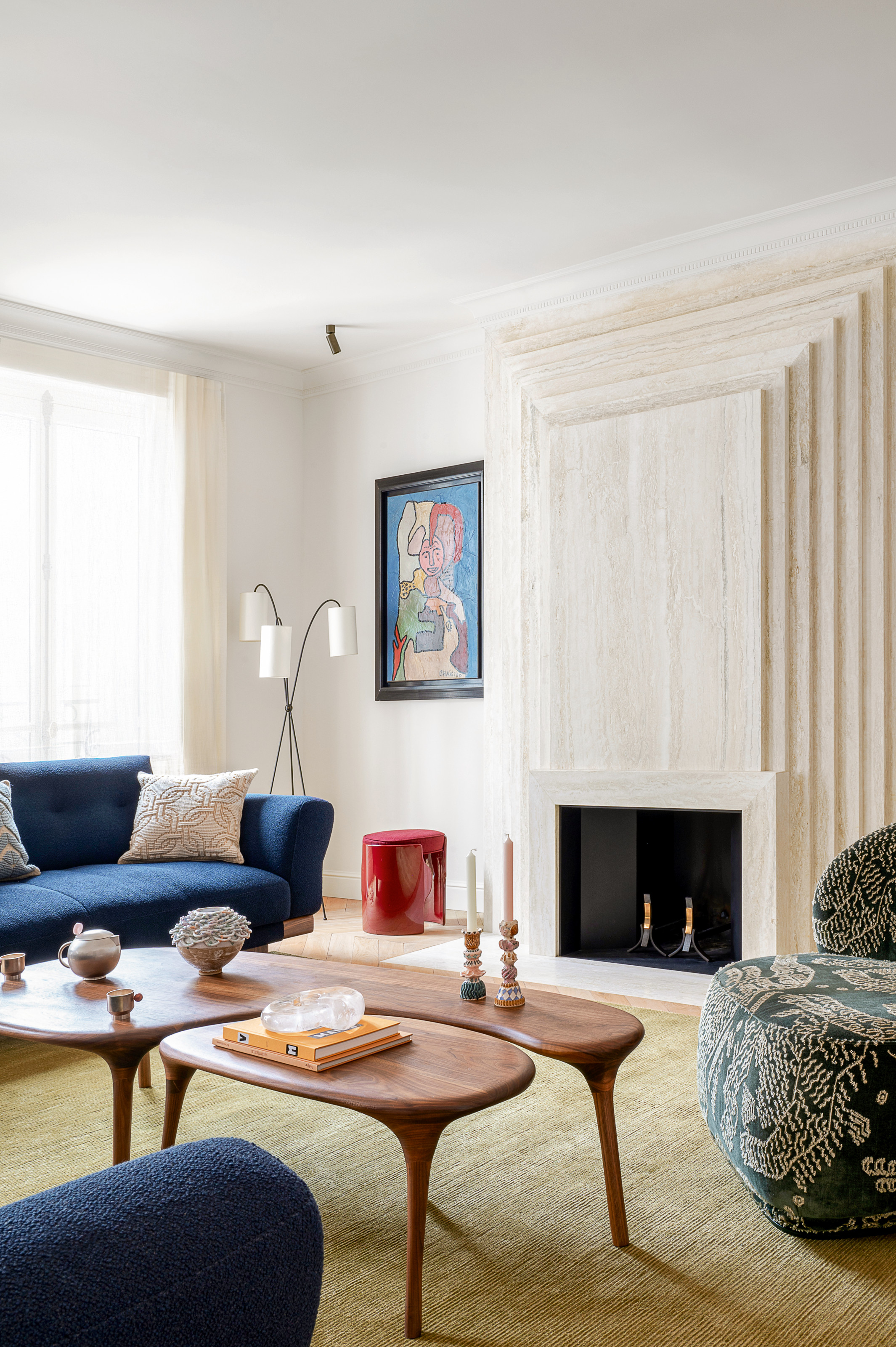 White living room with stone fireplace, pale green rug, dark blue sofa and sculptural wood coffee tables
