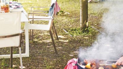 Person grilling on a barbecue outdoors with a white table and chairs in the background