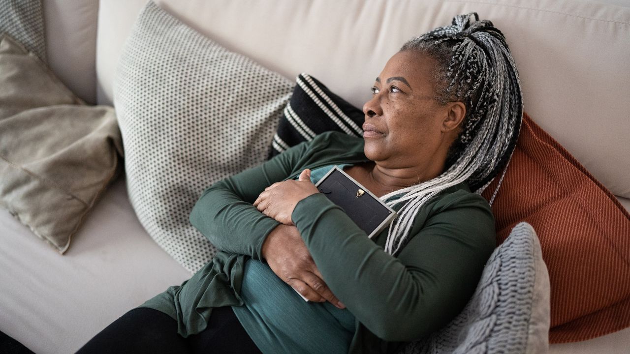 An older woman clutches a photo to her chest while lying on the sofa and looking out the window.