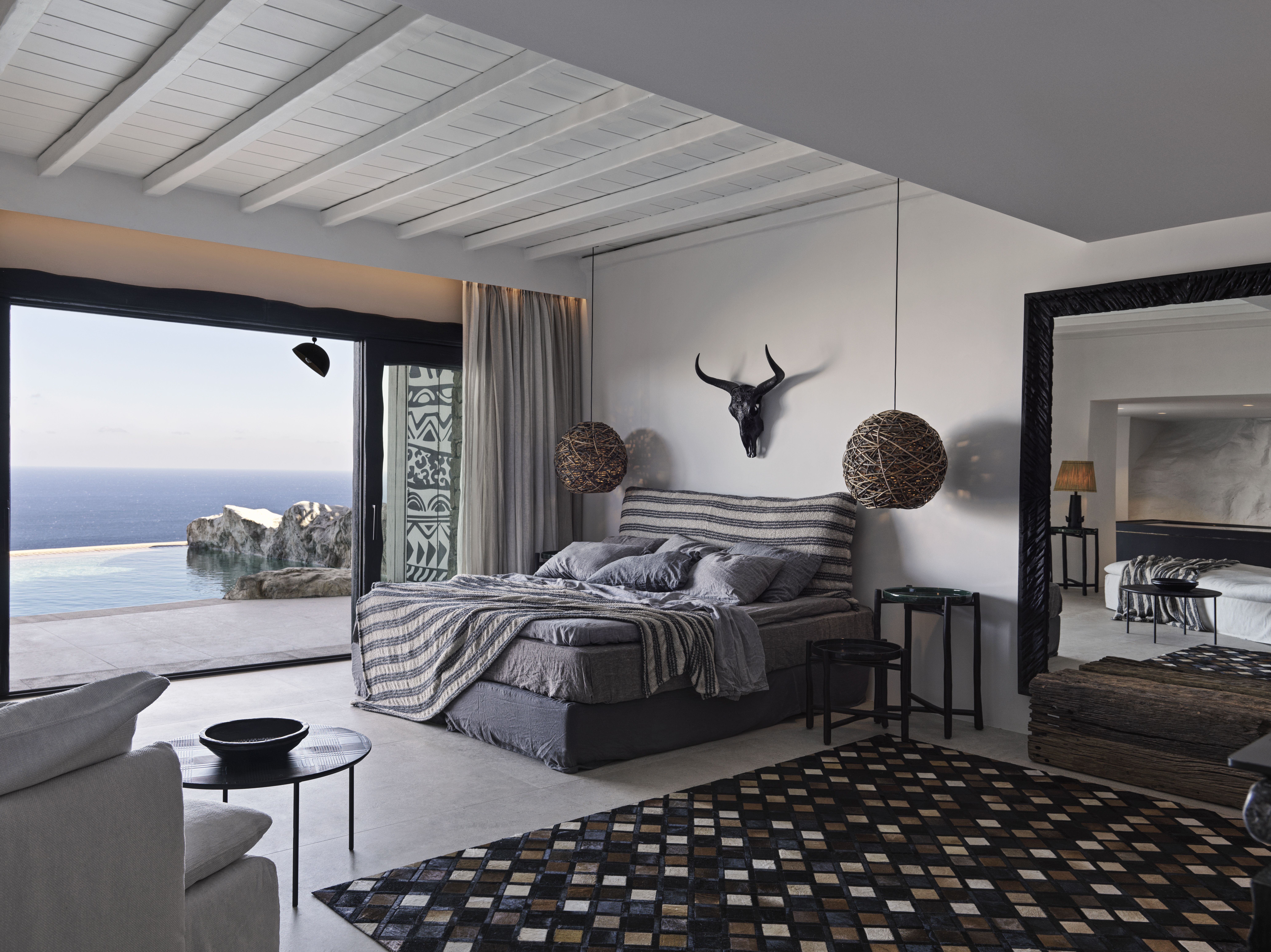 A black, beige, and white-tinted hotel room filled with animal skulls, rattan mirrors, stripy sheets, and seaside views.