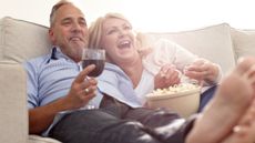 Mature mixed-sex couple relaxing together on the sofa with popcorn and wine, enjoying watching a movie