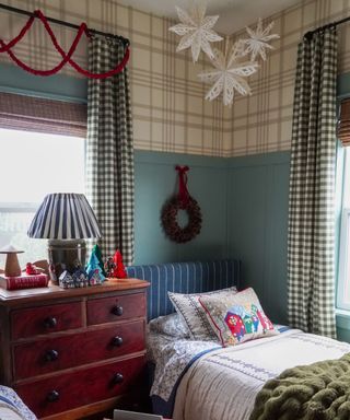 A kid's bedroom decorated with vintage Christmas decor featuring a wreath hung above the headboard, a red swagged garland on the curtain pole, white 3d paper snowflakes hung from the ceiling and tree and house ornaments on the bedside table