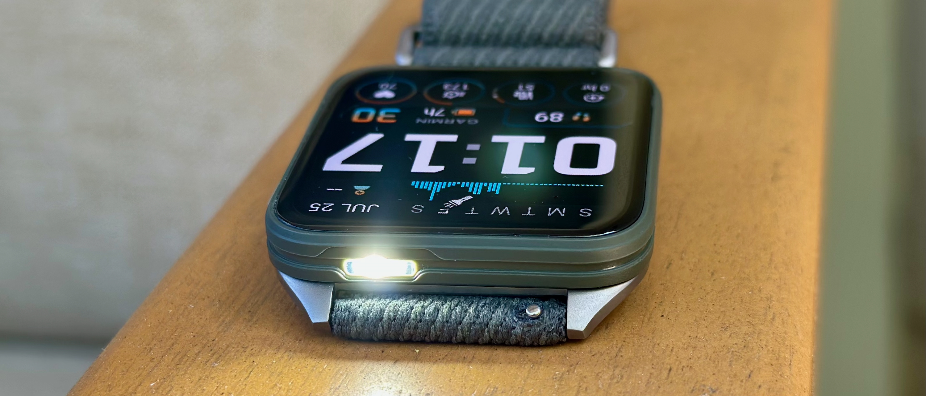 A photo of the Garmin Venu X1 sitting on an armrest, display up and facing opposite with the default watch face, the white LED flashlight on and pointing toward the camera.
