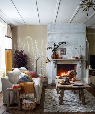 Living room with wooden floor and rug, grey sofa dressed with coloured cushions beside a wooden coffee table and lit fire.