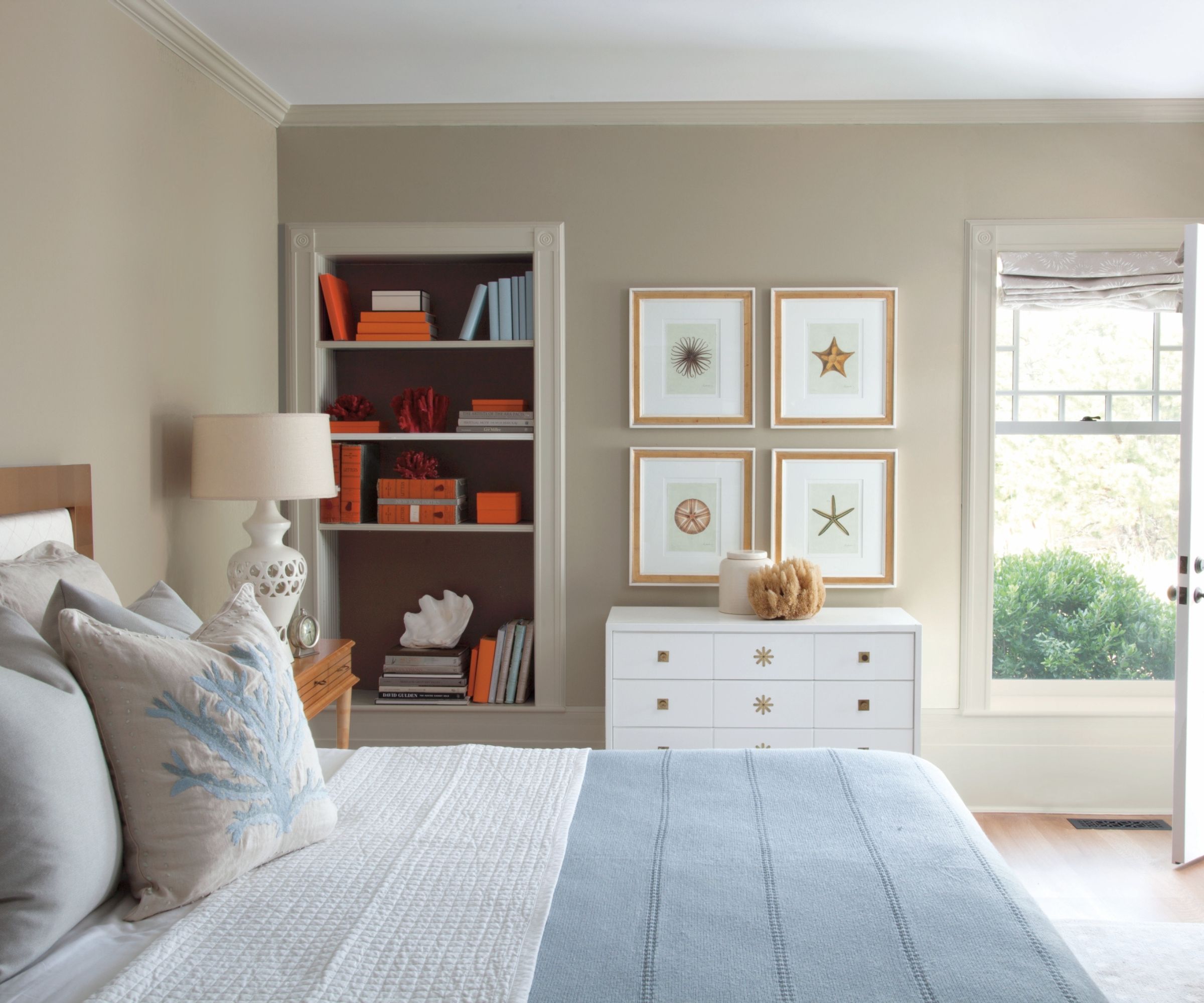 Bedroom with Benjamin Moore's Pashmina painted walls, bed with pale blue throw and nautical throw pillows, a white chest of drawers with coastal prints above and shelves with orange boxes and books
