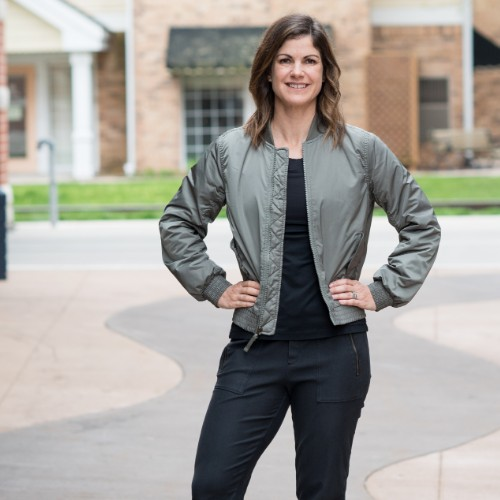woman facing the camera smiling with hands on her hips wearing a khaki jacket and black top and trousers. 