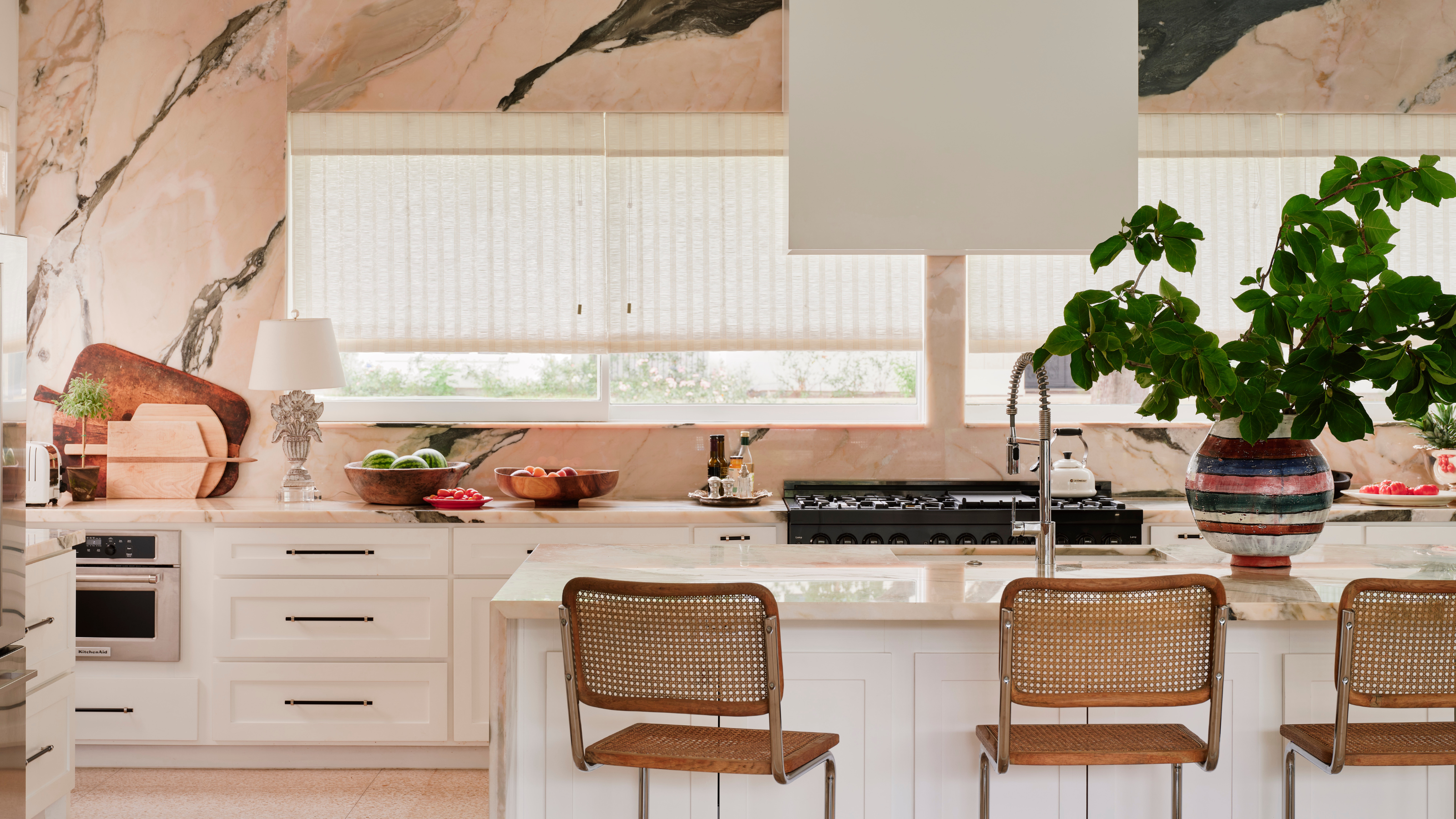 A white kitchen with a pink marble wall and smooth white cooker hood facing out large, wide windows opposite to the island.