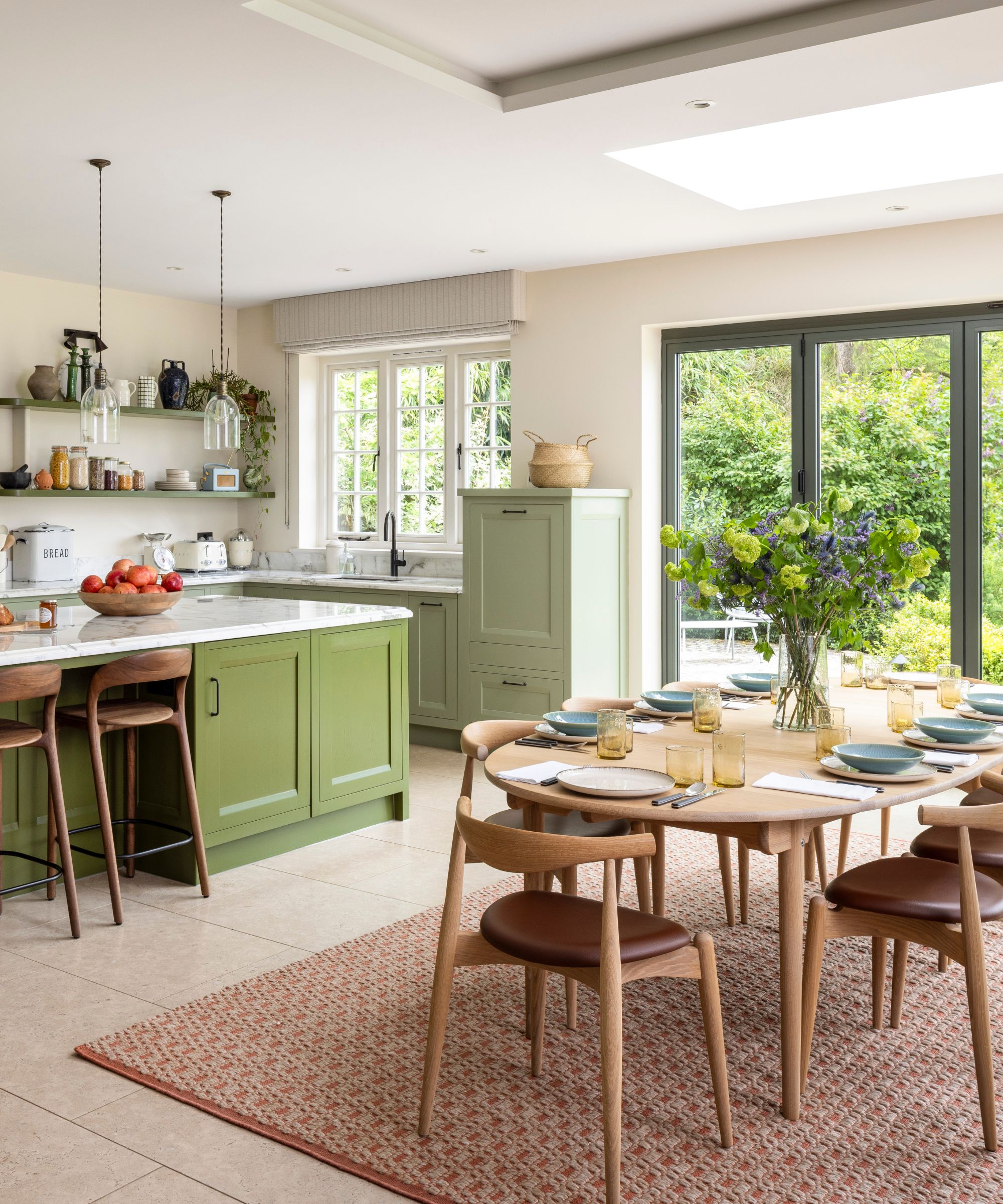 a green toned kitchen with neutral walls, large island, patio doors onto the garden, and a scandi style pale wood dining table and chairs