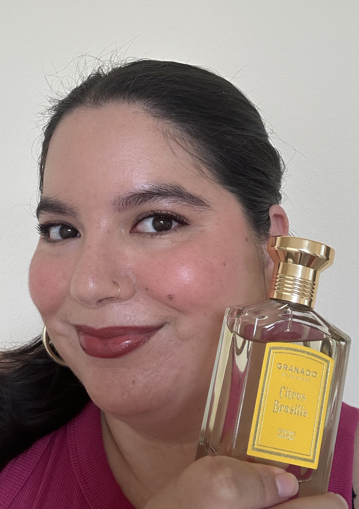 Holding up Granado Citrus Brasilis perfume next to my face, gold cap and bright yellow label with a glossy lip and rosy cheeks