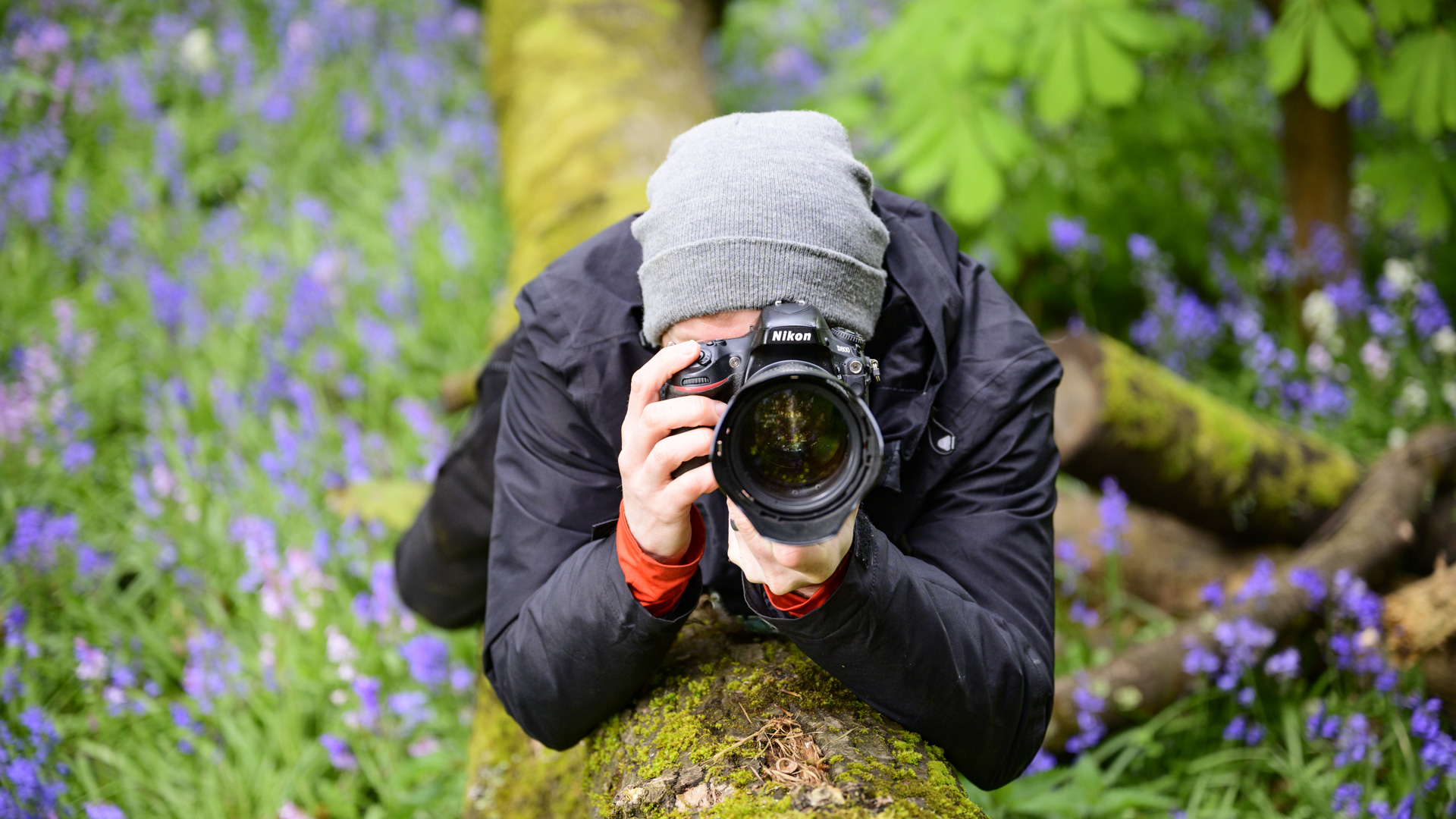 Photographer on a tree trunk surrounded by bluebells holding a Nikon D800 DSLR camera