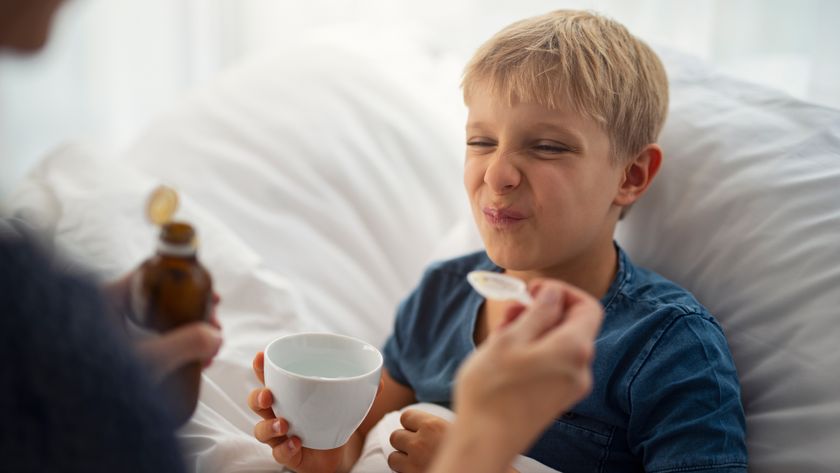 Sick little boy lying in bed making a face at his mother. The mother is giving a spoon of medicine to the boy. The boy hates the medicine and makes a disgusted face.