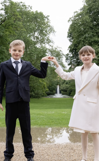 Prince Jacques wearing a suit and Princess Gabriella in a pink coat making heart hands in front of a grassy field and fountain