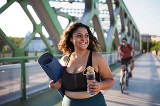 Mixed-race woman doing sport in city, carrying mat and water bottle.
