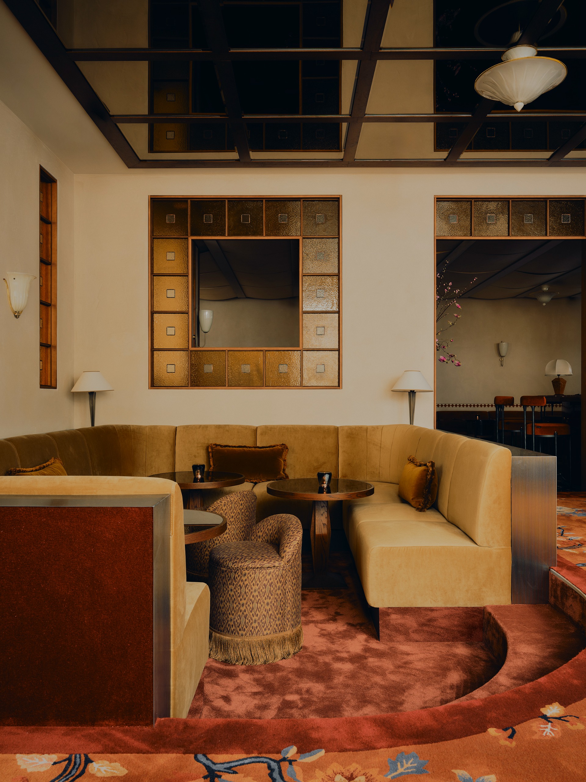 A modern conversation pit in a bar with stained glass windows, a mirror paneled ceiling and a floral carpet with trim, with ochre seating and brown throw pillows with small round bar tables and poufs