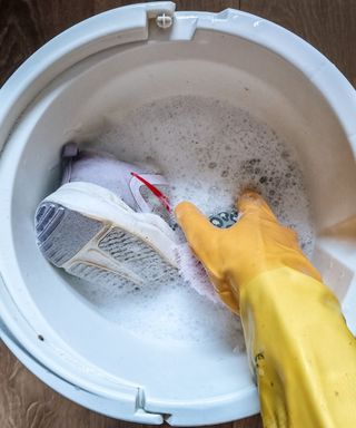 A birds eye view of a white bucket with soapy water, with a yellow marigold gloved hand cleaning a purple trainer with white soles