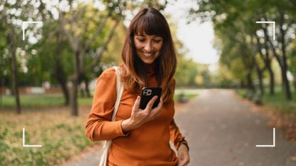 Woman with long hair standing in a park with trees behind her, looking at the best habit tracker apps on a smartphone and smiling